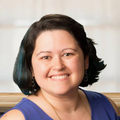 A woman with short dark hair, wearing a blue sleeveless top and dangling earrings, smiles at the camera against a blurred light background.