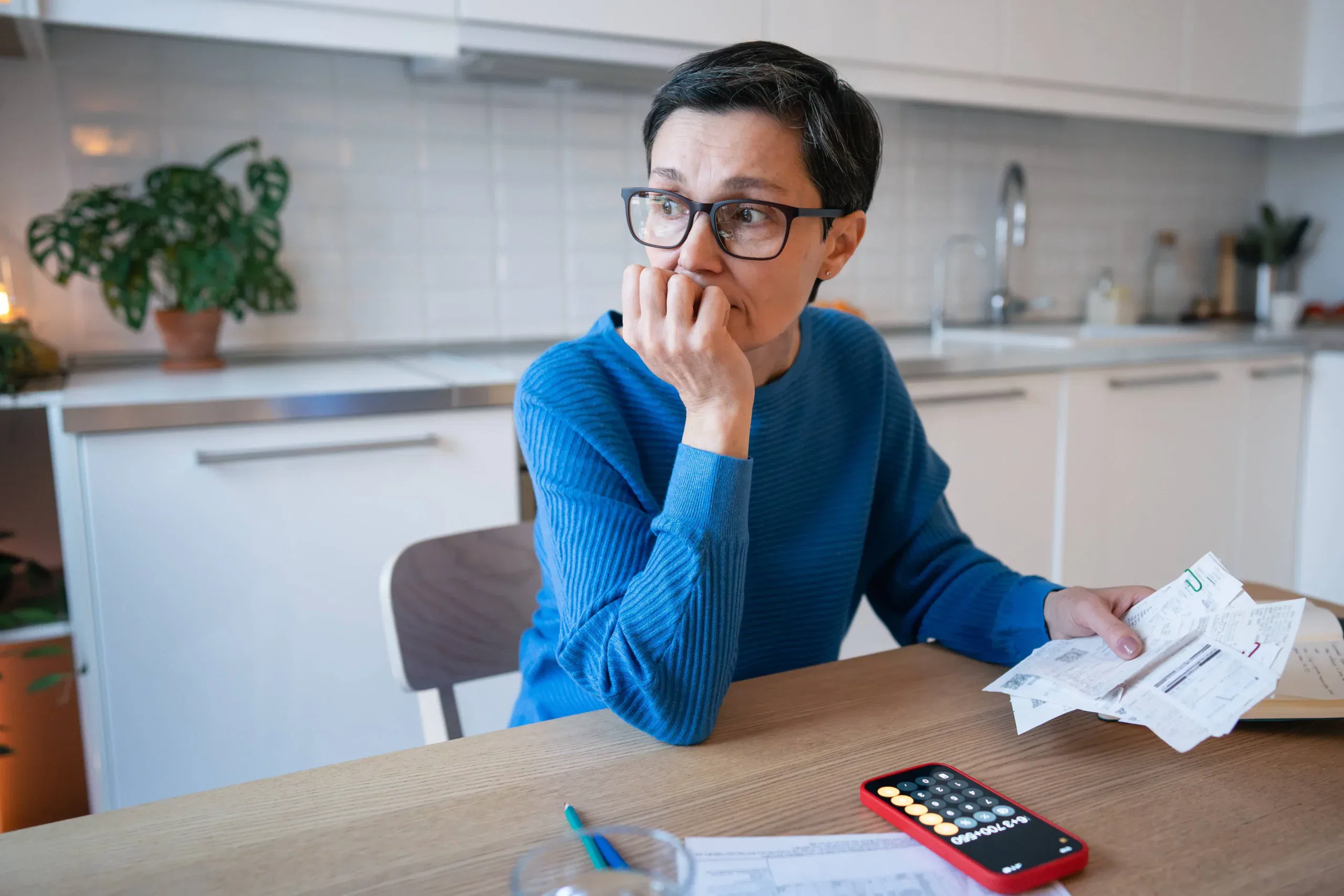 A person in glasses sits at a kitchen table holding bills, with a calculator app open on a smartphone and papers spread out, appearing concerned.